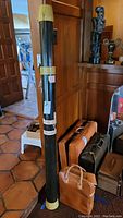 Tall green cylindrical Rod Guard fishing rod case with cream-colored caps, positioned vertically indoors against a wooden surface with cases and collectibles in background.