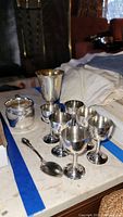 Overview of silver creamer, six goblet-style silver cups, cherry wood cups, and small silver spoon displayed on table.