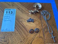 Photo showing six jewelry pieces on a wooden surface: an angel pin, a round textured brooch, two pairs of earrings (one with a colored circular center, the other clip-on style), and a silver chain with a cross pendant.
