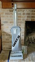 Front view of Oreck XL Classic vacuum cleaner upright showing cloth bag with ORECK branding, base unit and power cord against a brick fireplace background.