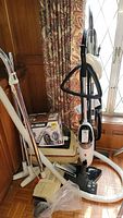 Photo showing multiple vacuum cleaners including Black and Decker boxed new model, Kenmore vintage upright vacuum, hoses and cleaning attachments.