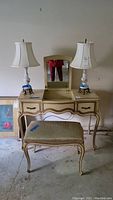 Full view of cream and gold French Provincial vanity with mirror and matching bench, flanked by two table lamps.