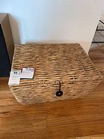 Photo of rectangular woven basket with a lid and handle, natural light brown color, placed on wooden floor with measurement tags visible.