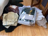 Photo showing variety of folded linens and bedding stack including fitted sheet package, patterned fabric pieces, blankets, and mattress pad.