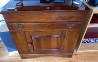 Front view of the wooden cabinet showing the drawer with two decorative metal handles and the single door with a round wooden knob and panel detailing.