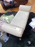 Light beige fabric chaise bench with curved backrest and wooden legs shown from a top angle on a black rug, surrounded by cushions and other furniture.