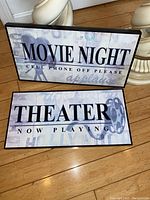 Two rectangular wall decor signs, one saying 'MOVIE NIGHT CELL PHONE OFF PLEASE' with a clapperboard, the other saying 'THEATER NOW PLAYING' with a film reel.