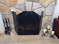 Black metal fireplace screen with decorative leaf cutouts in front of stone fireplace, set includes matching fireplace tools on stand and three candle holders with candles.
