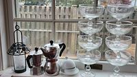 Wide shot of two metal stovetop espresso makers, white ceramic tea candle holder, stacked clear wide mouth glasses, and black metal lantern candle holder by window