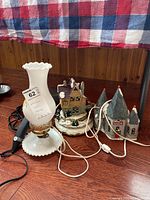 Photo showing milk glass hurricane lamp, Christmas houses, and black handheld massager on wooden table.