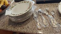 Stack of eight Christmas-themed China Pearl plates and four Lenox Holiday serving flatware pieces in plastic wrapping on countertop