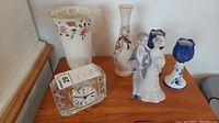 Photo showing entire lot with Lladro figurine, Lenox floral vases, Royal Doulton glass clock, and Delft Blue candle holders arranged on wood surface.