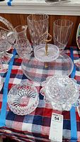 Photo showing various glassware items including two-tier serving platter, pitcher, vases, and candy dishes arranged on checkered cloth