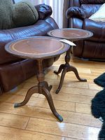 Front and side view of two wooden round side tables with painted circular ornamental design on the top, each with tripod pedestal base ending with metallic caps on legs. Positioned in front of brown leather seating on wooden floor.
