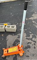 Orange hydraulic floor jack with a long silver handle on asphalt surface and two cinder blocks in the background.
