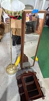 Seven wooden framed mirrors stacked leaning upright against a chair. A wooden wall-mounted display cabinet with glass doors is leaning in front. A tall floor lamp with a cream scalloped glass shade and brass base stands beside them.