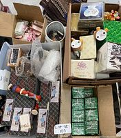 Top view of several cardboard boxes filled with assorted flea market items including wrapped glasses, small boxed collectibles, novelty mugs, and figurines, showing a variety of product packaging including Enesco boxes and personalized mug packaging.