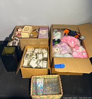 Two cardboard boxes containing assorted plush bears, boxed bear collectibles, and a basket with small magnets or items.