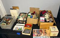 Wide view of multiple boxes and bins filled with assorted Christmas decorations including greenery, lights, trays, and figurines on a black tablecloth.