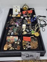 Top view of a black tray filled with assorted costume jewelry including chains, pendants, brooches, earrings and bracelets in mixed metals and colors.
