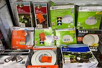 Photo showing multiple boxed LED lights including Hampton Bay flood lights, Commercial Electric LED flush mounts, and recessed lights, plus Defiant LED security light.