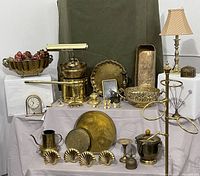 Full view of all brass items displayed on a table with beige cloth backdrop showing towel ring stand, lamp, clock, bowls, trays, napkin rings, incense box, candle holder, and more.