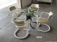 Four white cantilever chairs arranged around square glass-topped table with black metal base. The chairs have tubular steel frames and creamy white vinyl seats. Table glass shows minor chip.