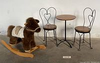 Photo showing entire lot: child-sized bistro table and two chairs next to plush rocking horse with wooden rockers.