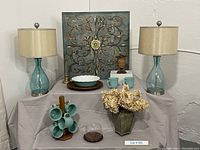 Pair of matching teal glass lamps with beige shades, teal ceramic mugs on wooden mug tree, canvas wall art, metal vase with dried flowers, ceramic pie plate, wooden lazy susan visible.