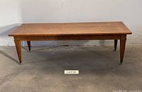 Front view of midcentury modern hardwood coffee table with tapered legs and casters shown on a concrete floor against a white wall.