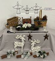 Photo showing the assembled collection on gray cloth: multiple woven baskets, pinecones, wire angels on wall, white wooden reindeer, cinnamon bundles with floral accents, metal star ornaments on strap, jingle bells, and candle holders.