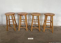 Four wooden stools made of light hardwood arranged in a row against a plain wall on concrete floor. Each stool has a round top and four legs linked by horizontal supports. Tops show scratches and wear.