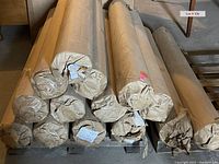 Twelve rolls of wallpaper stacked on a pallet, wrapped in brown paper with tags visible on several rolls.