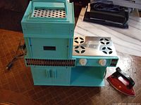 Full view of the turquoise 1958 Kenner Easy Bake Oven next to the small red and black electric iron on a wooden surface.