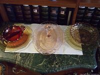 Seven pieces of antique Depression glass displayed on a green marble countertop with a white cloth underneath. Includes various plates, bowls, and a server with handle. Visible red bowl with silver overlay.