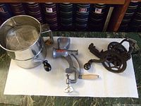 Photo showing four antique kitchen gadgets on a white paper towel in front of books. Flour sifter is on left, nutmeg grinder center-left, apple peeler black cast iron with gears center-right, coffee grinder on right.