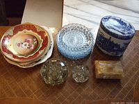 Overview of stacked decorative plates, clear flower frogs, blue and white lidded jar, onyx trinket box, and assorted items on patterned surface.