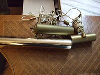 Two older picture frame lights shown on a brown patterned surface. One light features shiny chrome finish and the other has a brass finish. Each has attached power cords and mounting hardware.