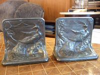 Pair of cast metal bookends with embossed crane bird design showing front faces on a checkered surface with wood-paneled background.