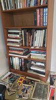 Wooden bookshelf with three shelves housing an assortment of hardcover and softcover books, plus loose items on floor
