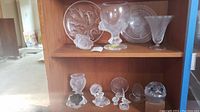 Two-tier wooden display with multiple Lalique crystal pieces including plate, goblet, vase, dish on top shelf and small figurines and paperweights on bottom shelf