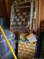 Five metal framed lawn chairs with woven webbing seats and backs, leaning against wall with wooden armrests visible. Yellow and white striped plastic cover and packaging for chaise lounge visible at bottom.