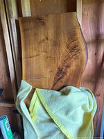 Live edge wood slab leaning vertically against a wooden wall, showing polished top surface with natural cracks, a yellow blanket partially covering the lower part, visible scratches, and a natural edge with bark.