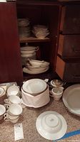 Noritake china plates, cups, and covered tureens arranged inside a wood cabinet and on a carpeted floor. Cups stacked in front, plates and other serving pieces stacked inside the cabinet shelves.