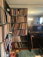 Metal shelving unit with six shelves filled with vinyl record albums; upright piano and cabaret cane leaning against it
