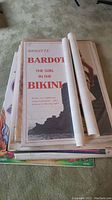 Stack of rolled and unrolled vintage posters on carpet, top poster Brigitte Bardot “The Girl in the Bikini” partially rolled