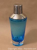 Full view of blue art glass cocktail shaker with stainless steel top against beige background.