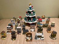 Entire lot showing one ceramic Christmas tree and 27 Baileys miniature collectible houses and ornaments arranged on a wooden table.
