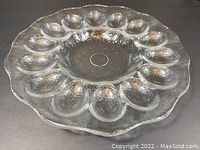 Clear textured glass egg-serving dish shown from above on a dark background, displaying 14 egg-sized depressions around a smooth center with scalloped edges.