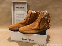 Side view of the Minnetonka Two-Button boots on a box, showing one full boot side profile with fringe and concho buttons, tan suede color.